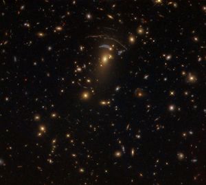 Black background dotted with galaxies. A reddish-orange galaxy at image center with a bluish-white arc above it.